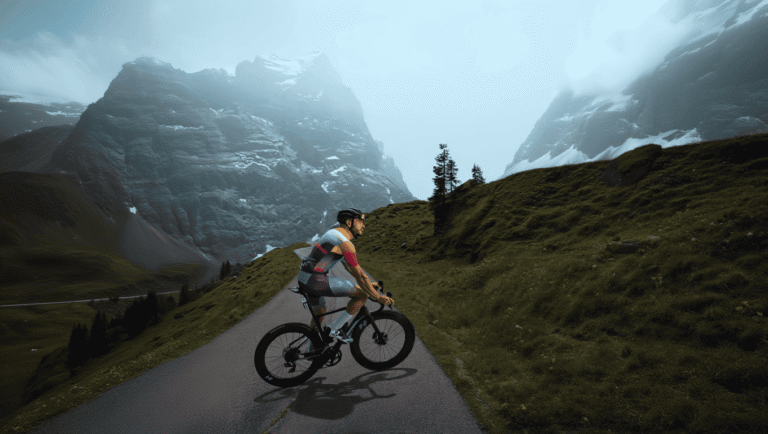 Ciclista pedalando em estrada montanhosa épica nos Alpes com picos nevados e neblina ao fundo, vibe de aventura no ciclismo MTB 2026