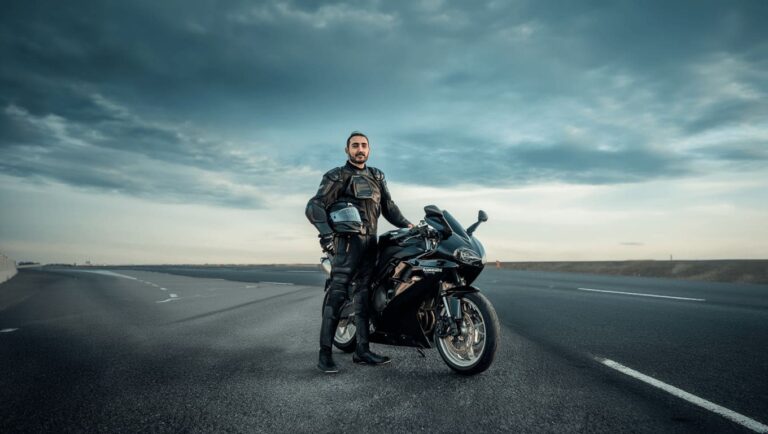 Motociclista com equipamento de proteção completo — capacete, jaqueta, luvas e calça — posando ao lado de moto esportiva em rodovia